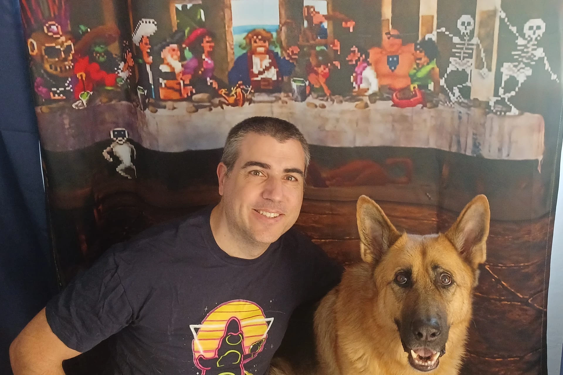 Miguel García sonríe junto a su pastor alemán, LeChuck, delante de una cortina con los personajes de Monkey Island en pixel art sentados como en la Última Cena