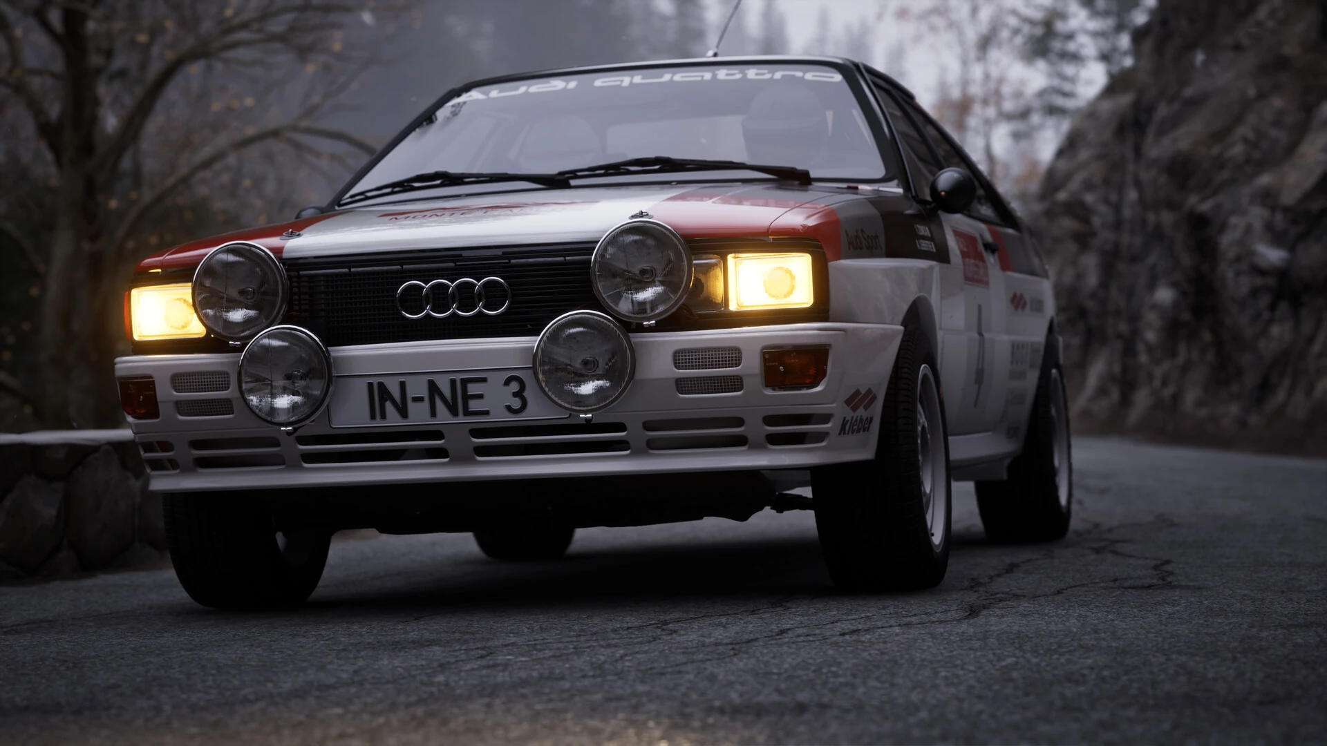 Un Audi Quattro blanco y rojo de rally, con cuatro faros auxiliares encendidos, en una carretera de montaña