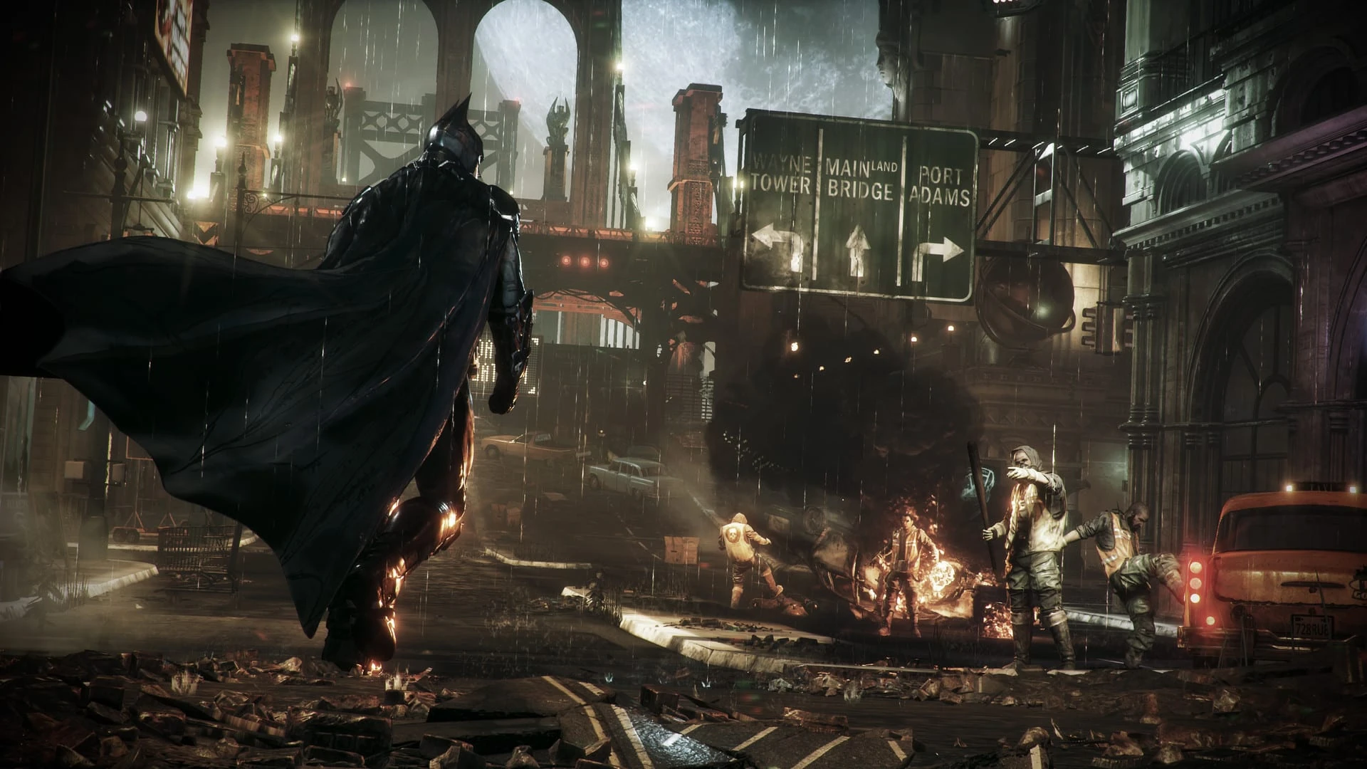 Batman, de espaldas y con la capa al viento, camina bajo la lluvia por una calle de Gotham con coches y escombros ardiendo y un grupo de matones al fondo