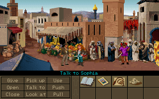 Indiana Jones y Sophia Hapgood en un zoco abarrotado; abajo, la rejilla de verbos del motor SCUMM y el inventario, con el cursor formando la orden Talk to Sophi