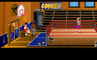Gimnasio universitario con un ring de boxeo a la derecha, un boxeador rubio dentro y Indy de pie junto a un banco, bajo un cartel que dice Cougars