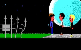 Tres chavales de pie sobre la gravilla ante la verja de la finca, de noche, con una luna enorme detrás