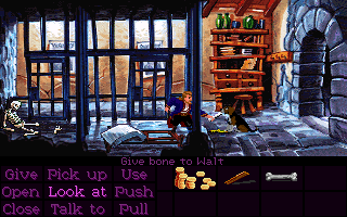 Guybrush Threepwood, de pelo rubio y chaqueta azul, agachado ante un perro pequeño en una cárcel de piedra; abajo, la rejilla de verbos de SCUMM y el texto Give
