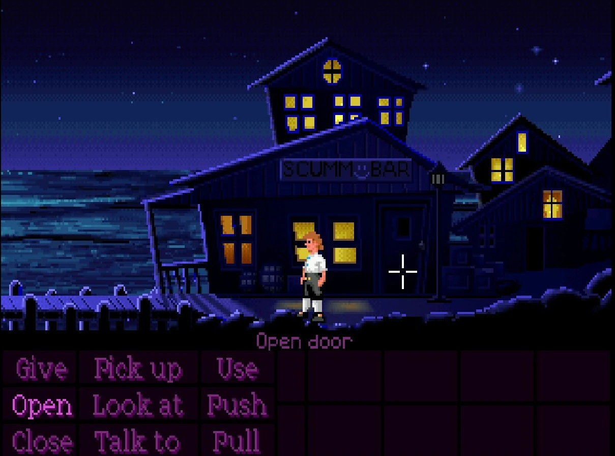 Guybrush Threepwood, de noche, delante de la puerta del SCUMM Bar en Mêlée Island, con la rejilla de verbos del motor SCUMM en la mitad inferior de la pantalla