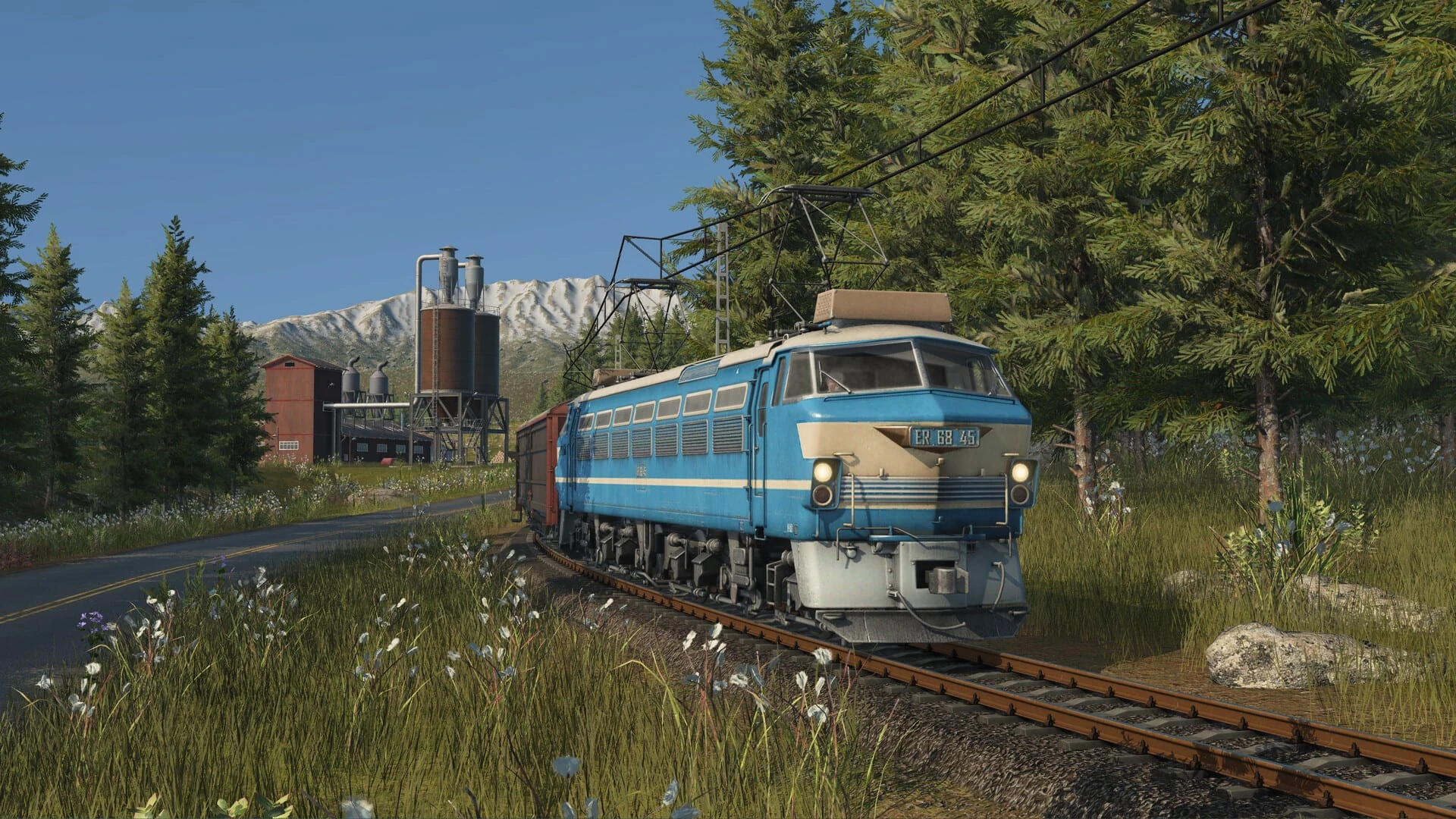 Una locomotora eléctrica azul y crema arrastra vagones de mercancías entre pinos, con una fábrica de silos y montañas nevadas al fondo