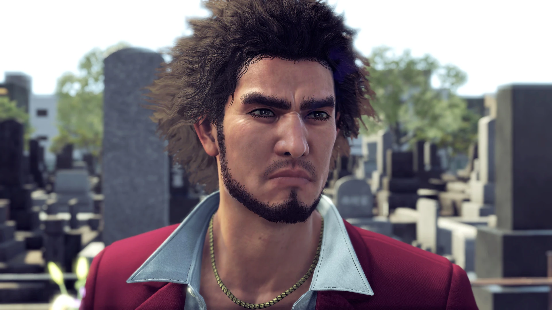 Ichiban Kasuga, con traje rojo y cadena al cuello, mira serio a cámara en un cementerio lleno de lápidas