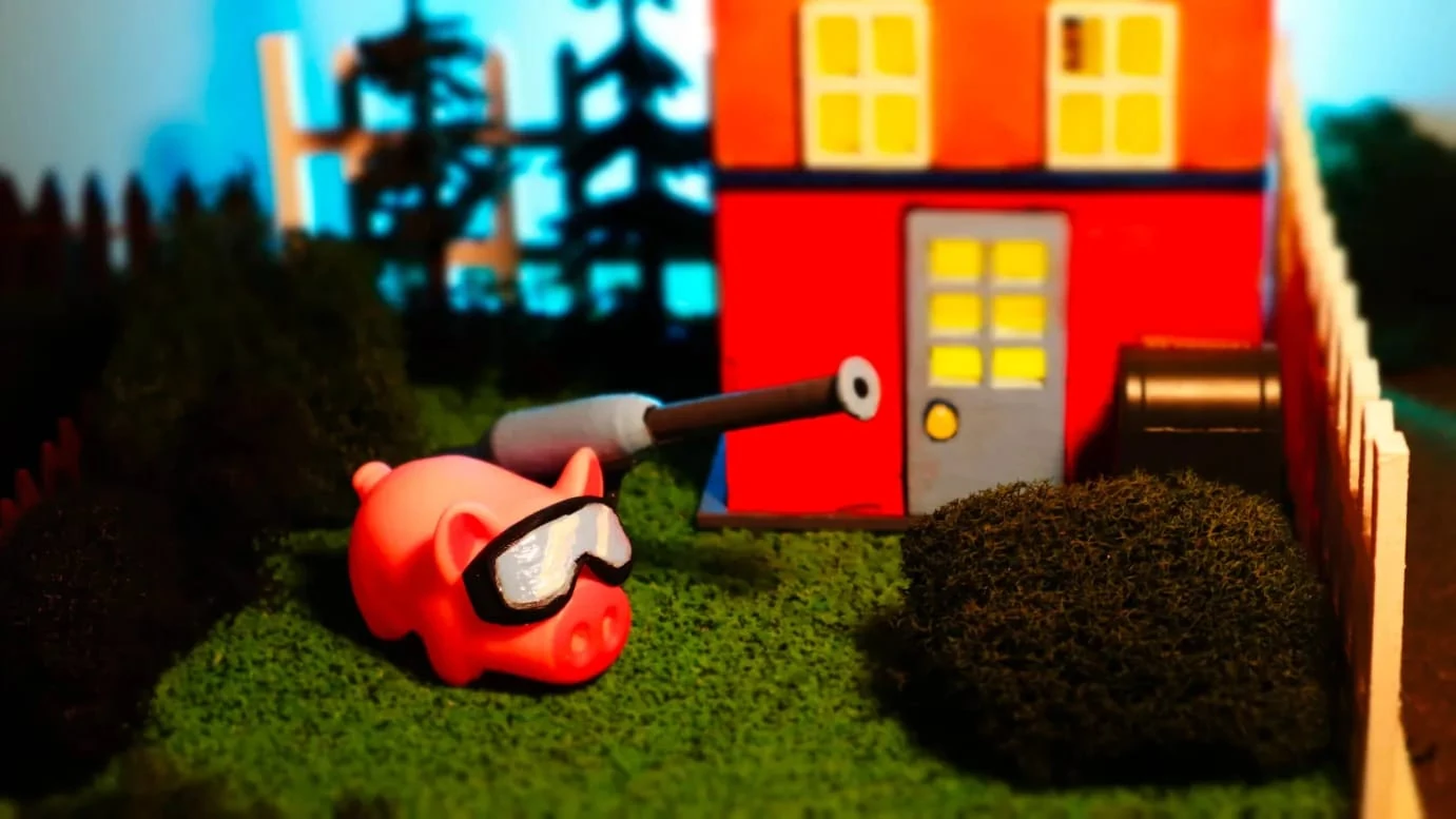 Una figura de Pigxel, un cerdo rosa con gafas de buceo, apunta con la lanza de una hidrolimpiadora a una casita roja de ventanas amarillas, junto a un seto