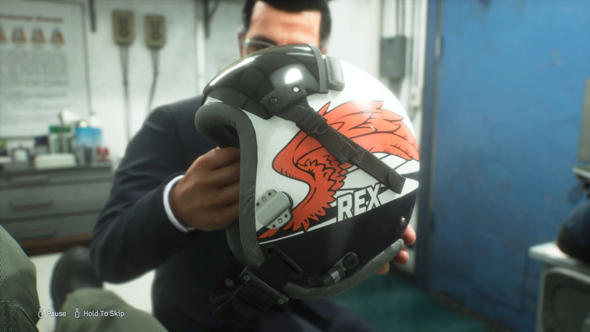 Un hombre con traje y gafas sostiene un casco de piloto blanco con unas alas rojas pintadas y el nombre Rex