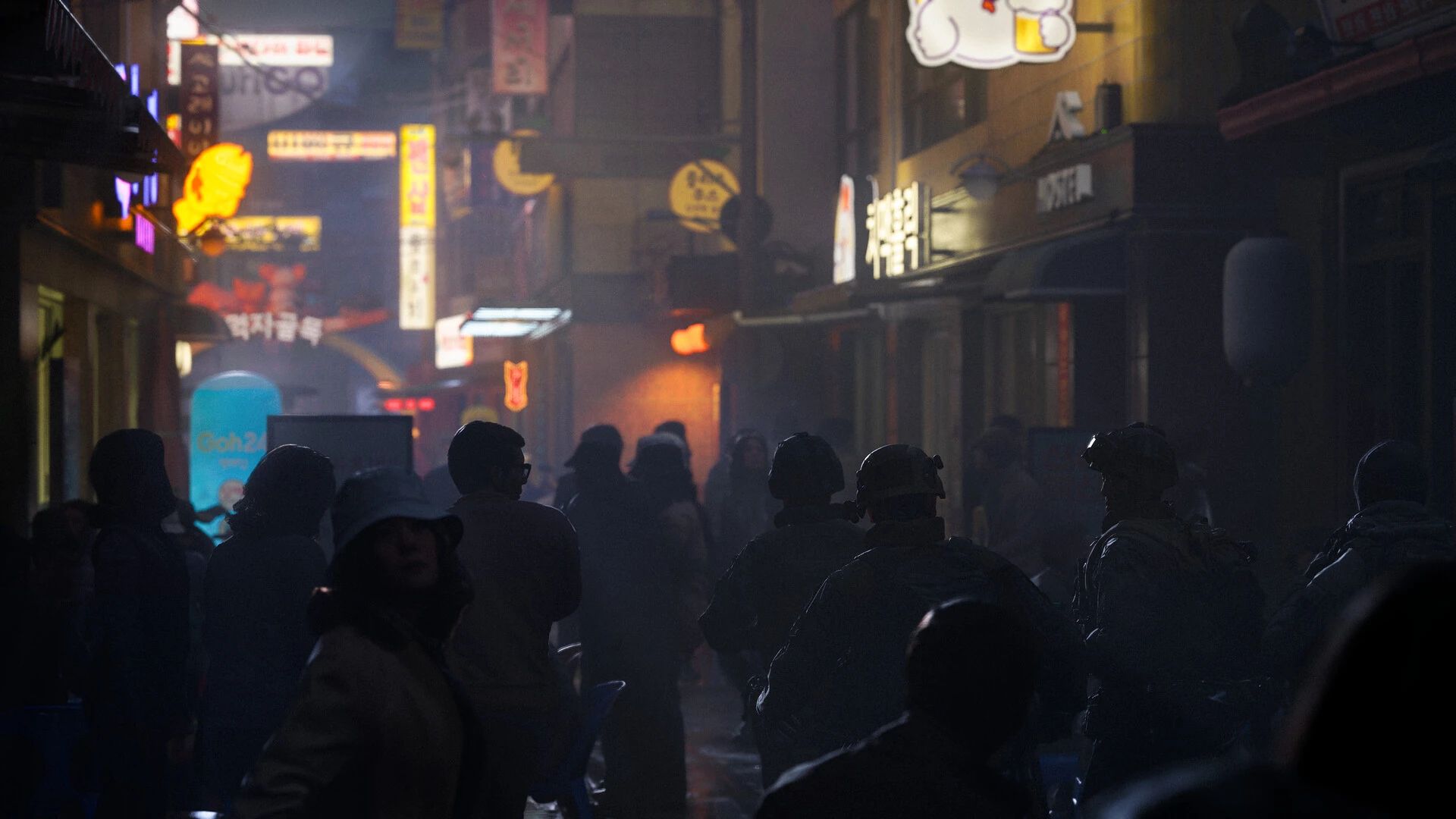Siluetas de soldados y civiles en un callejón estrecho lleno de letreros luminosos en coreano