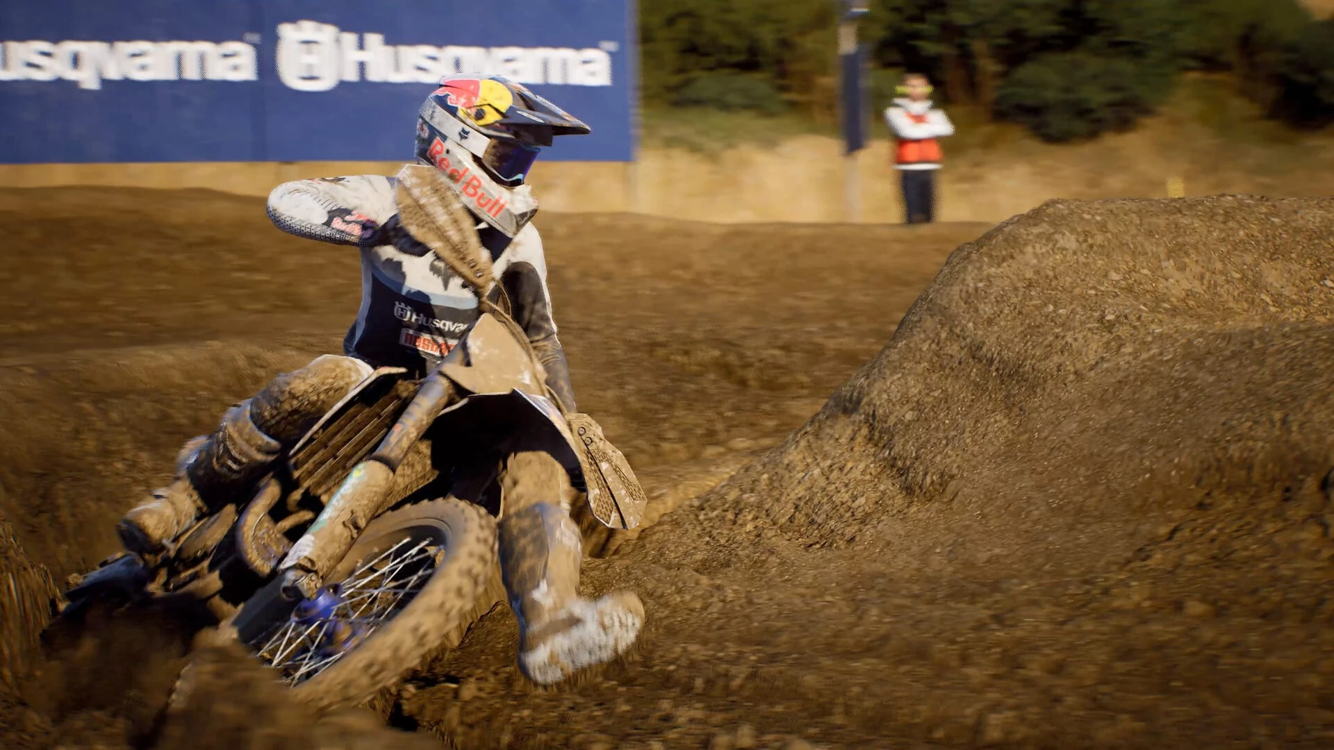 Un piloto cubierto de barro inclina la moto en una curva cerrada junto a un montículo de tierra, con un comisario al fondo