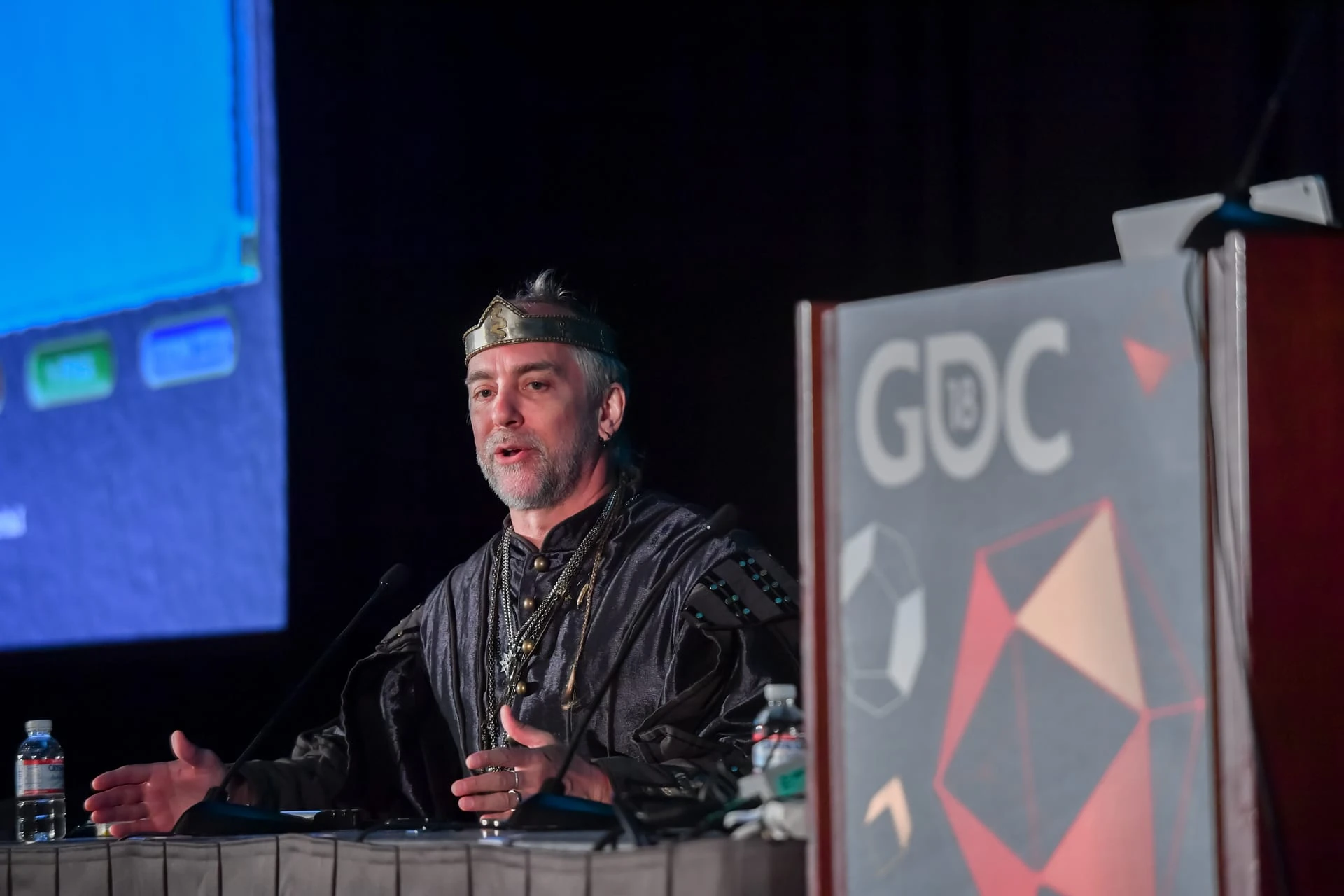 Richard Garriott, con una corona dorada y una túnica negra, habla ante un micrófono en un escenario de la GDC de 2018