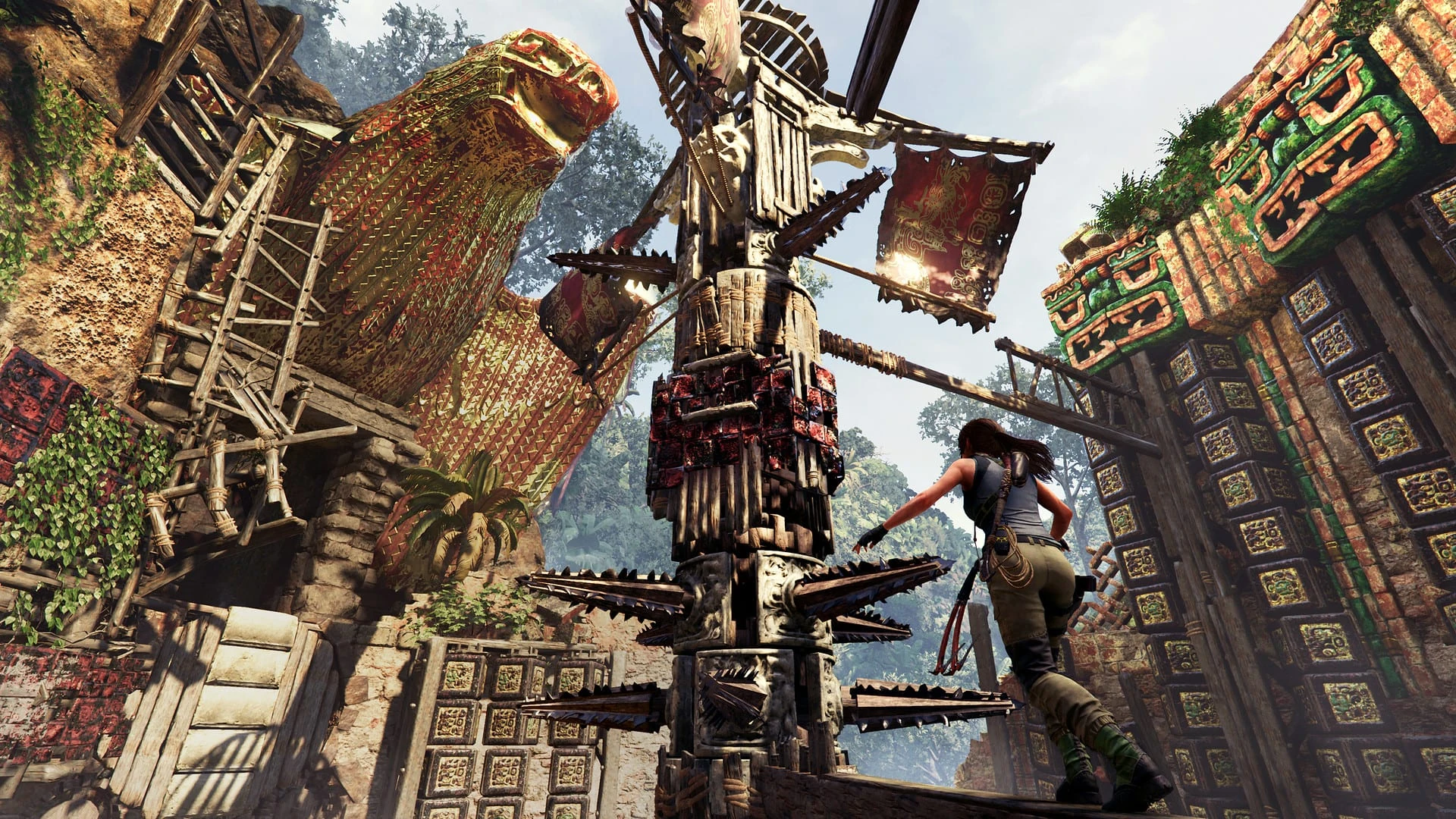 Lara Croft cruza una pasarela junto a una torre de madera con cuchillas giratorias, entre edificios de piedra pintados en la selva
