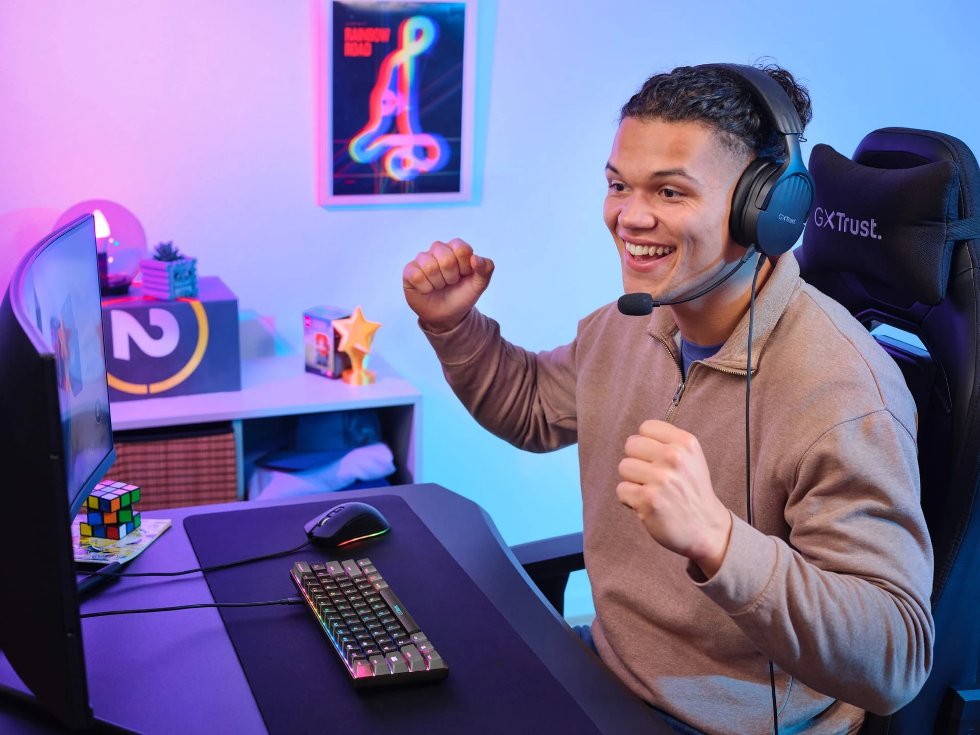 Un joven celebra con los puños en alto ante el monitor, con auriculares GXT, un teclado compacto con luces RGB y una silla gaming de GXTrust