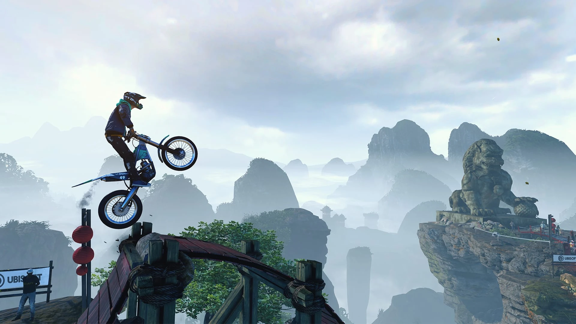 Un piloto de trial salta con su moto desde un puente de madera rojo sobre un paisaje de picos de piedra entre la niebla, con una estatua de león a la derecha